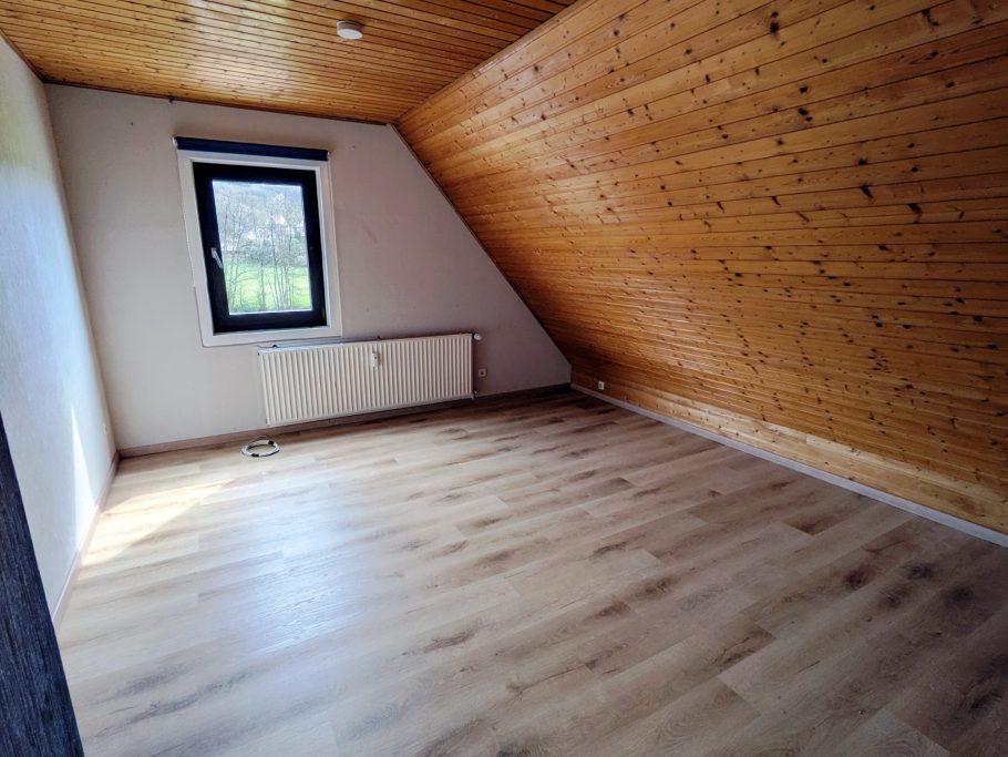 Räumlicher, unmöblierter Raum mit Holzdecke und Fenster. Helle Bodenfläche.