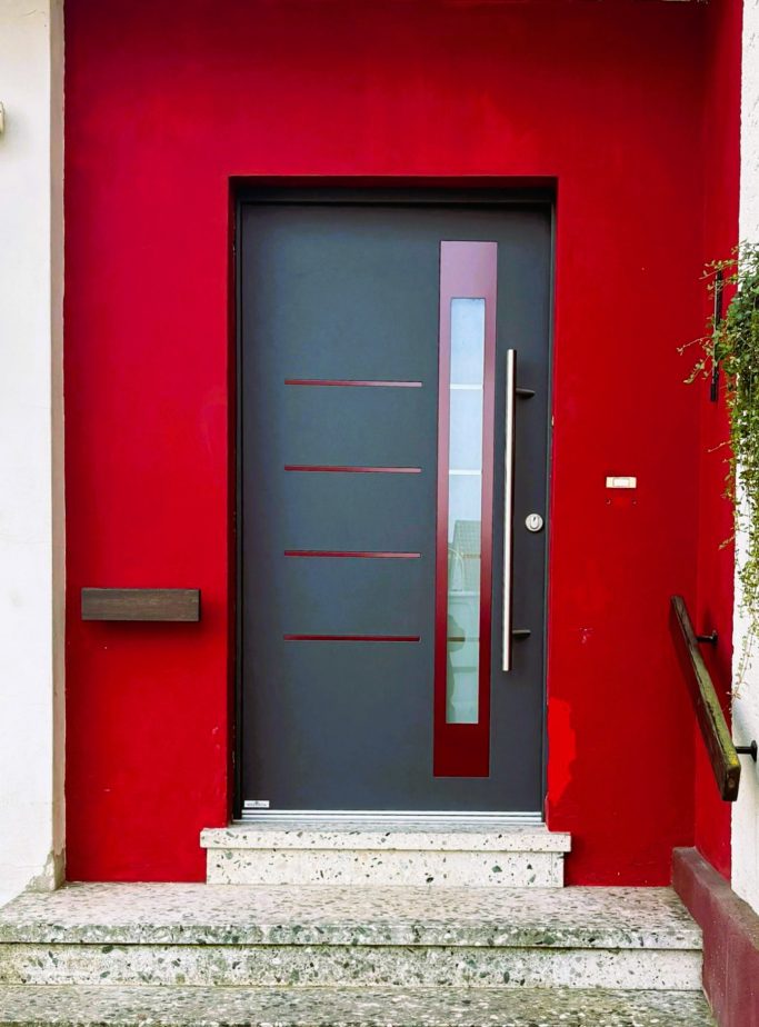Graue Haustür mit Glaselementen vor roter Wand und Treppe.